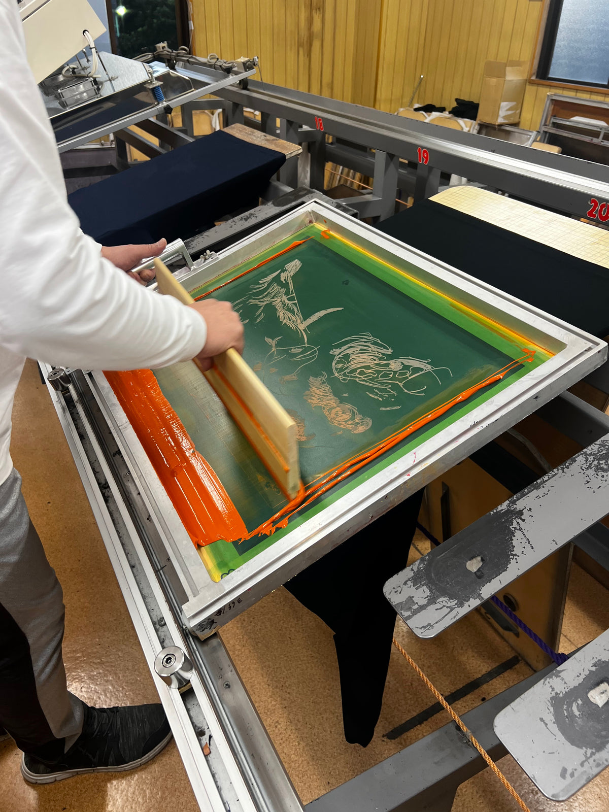 A person using a wooden screen printing squeegee to apply orange ink onto a green screen frame mounted on a large metal printing press in a workshop.